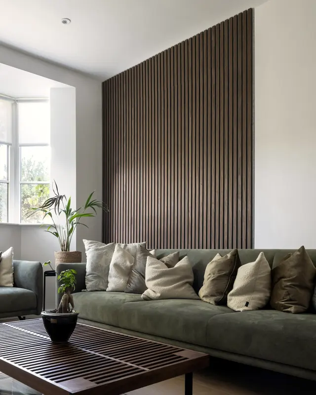 Wohnzimmer mit dunkler Holzlamellenwand hinter einem grünen Sofa und einem Lattentisch.