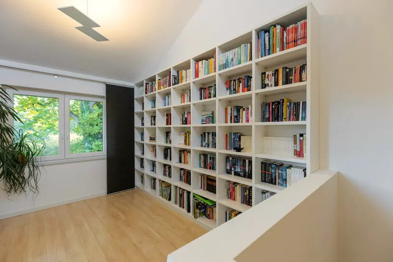 Weiße, raumhohe Bücherwand unter einer Dachschräge in einem hellen Flur mit Fenster.