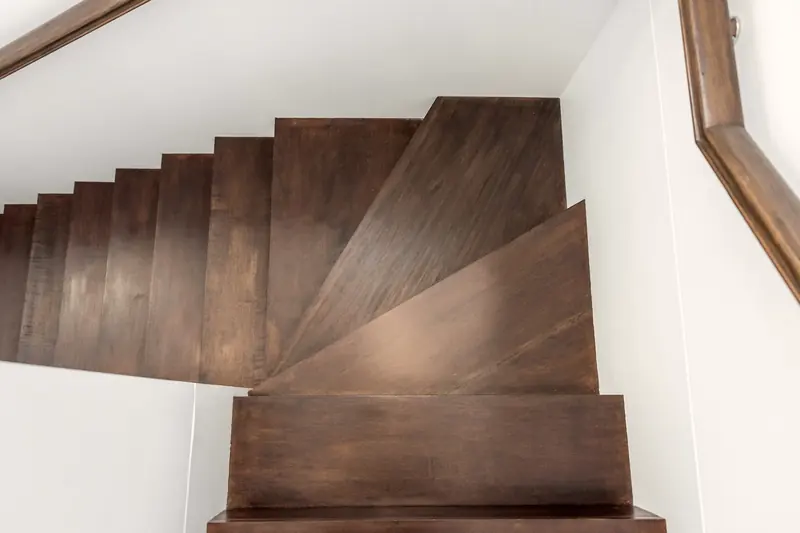 Viertelgewendelte Treppe mit dunkel geölten Holzstufen zwischen weißen Wänden, darüber ein Holzhandlauf.