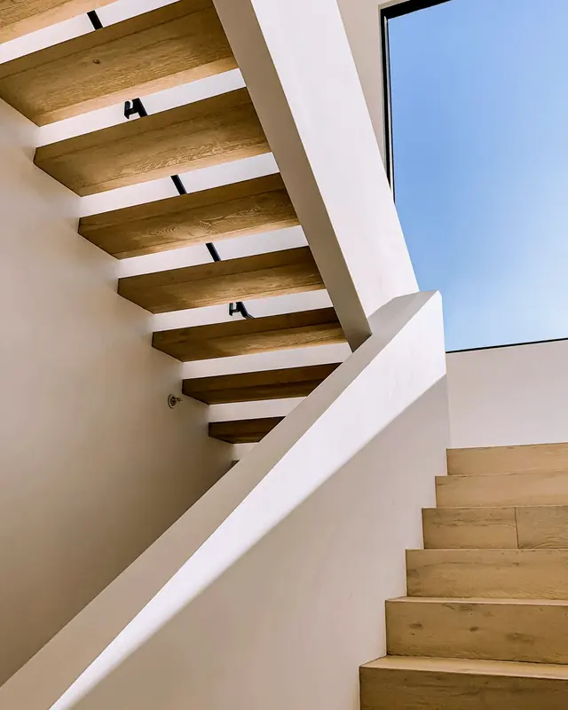 Blick von unten auf eine Kragarmtreppe: massive Eichenstufen ragen aus einer weißen Wand, oben ein Oberlicht mit blauem Himmel.