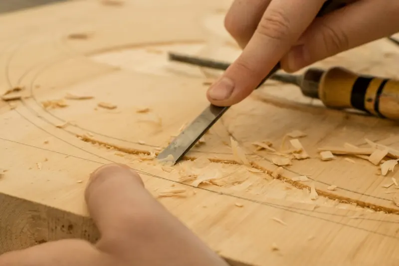 Hand führt einen Stechbeitel entlang einer angerissenen Kurve auf einem Holzbrett, daneben Späne.